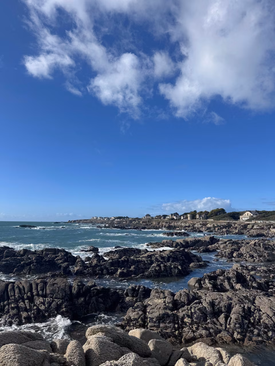 Côte rocheuse bretonne sous un ciel bleu parsemé de nuages blancs.