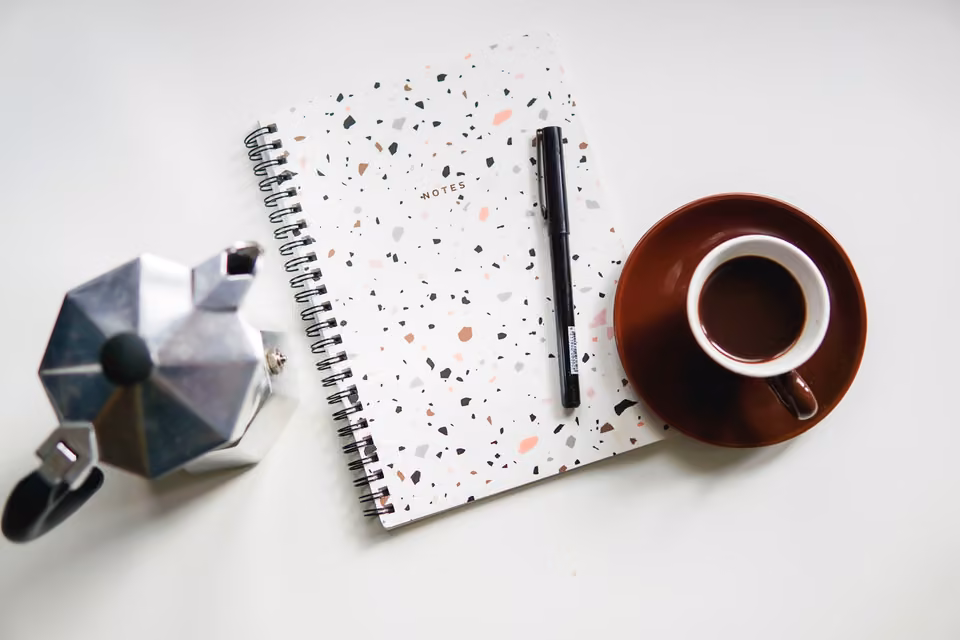 Carnet, stylo, café et cafetière italienne sur un bureau blanc.